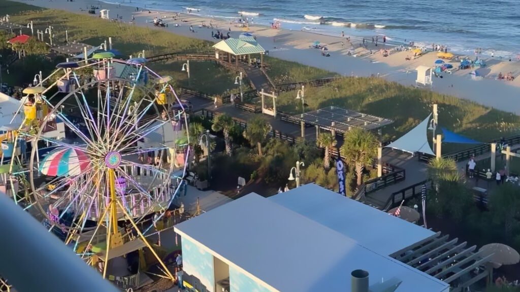 Vibrant high-angle view of a lively Carolina beach scene with a large, colorful Ferris wheel in the foreground, boardwalks, palm trees, and beach pavilions. A wide sandy beach stretches toward the ocean, filled with beachgoers, colorful umbrellas, and gentle waves under a sunny sky