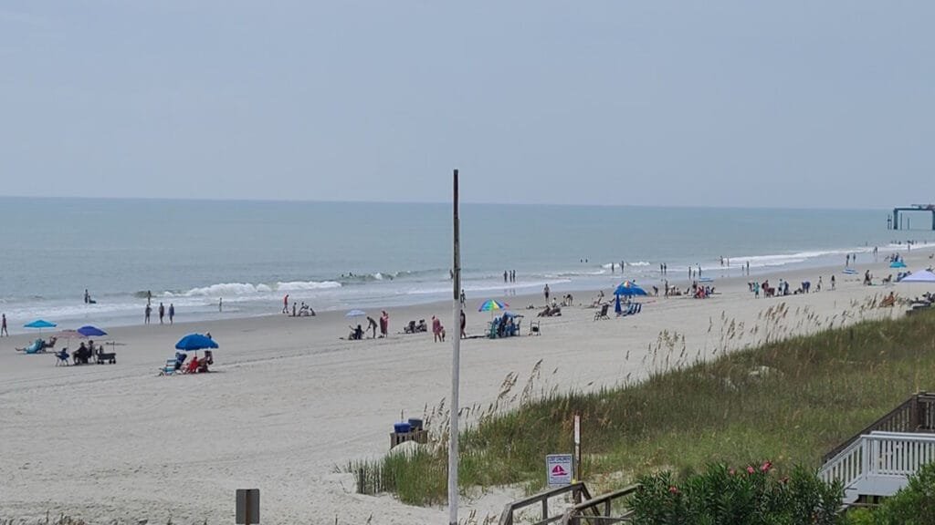 An wide shot of a sandy beach on an overcast day shows people relaxing under colorful umbrellas near the ocean waves, with a wooden staircase and beach grass in the foreground.