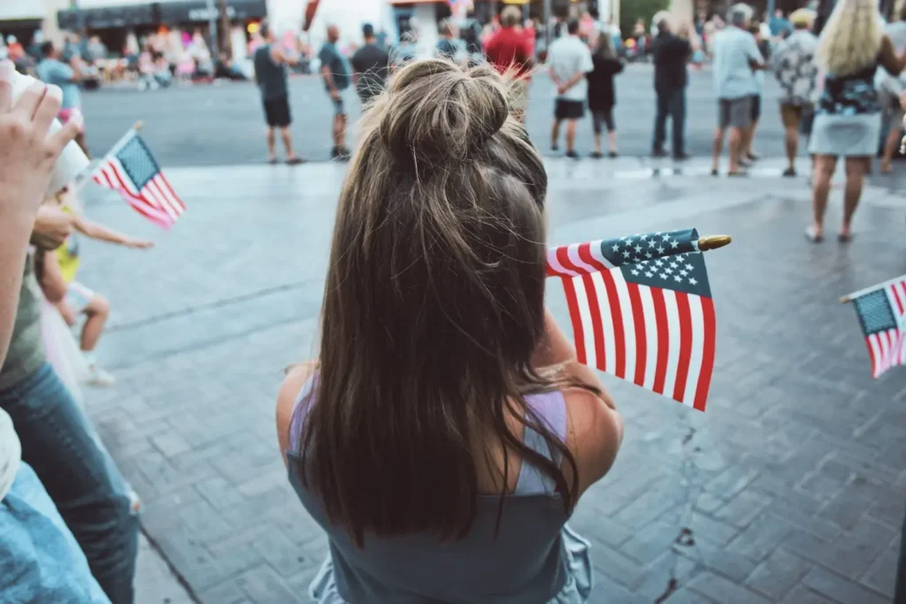 Viewed from behind, a young woman with long brown hair in a bun stands on a brick-paved street holding a small American flag. She is part of a crowd gathered outdoors, likely at a patriotic event or 4th of July celebration, with other people and American flags visible in the background.