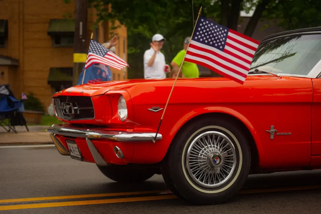 Bright red vintage Ford Mustang convertible decorated with American flags drives down a city street during a Fourth of July parade. Spectators wave from the sidewalk in the background as the classic car rolls past on an overcast day.