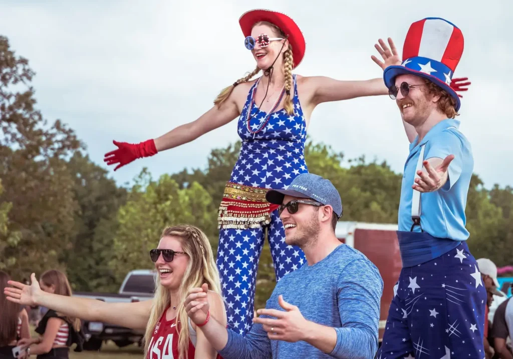 A group of smiling people dressed in patriotic red, white, and blue outfits celebrate outdoors during a Fourth of July event. Two participants wear star-spangled costumes and festive hats while others in sunglasses raise their arms and cheer against a backdrop of trees and parked vehicles.