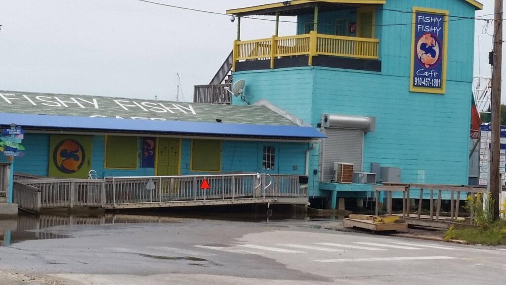 A wide view of the Fishy Fishy Cafe, a bright turquoise two-story waterfront building with yellow railings and accents. The lower section has a blue roof with large white 'FISHY FISHY CAFE' lettering. A tall vertical sign on the right tower shows the cafe’s name, a circular fish logo, and phone number 910-457-1881. A wooden ramp and deck extend over the water in front of the building under an overcast sky.