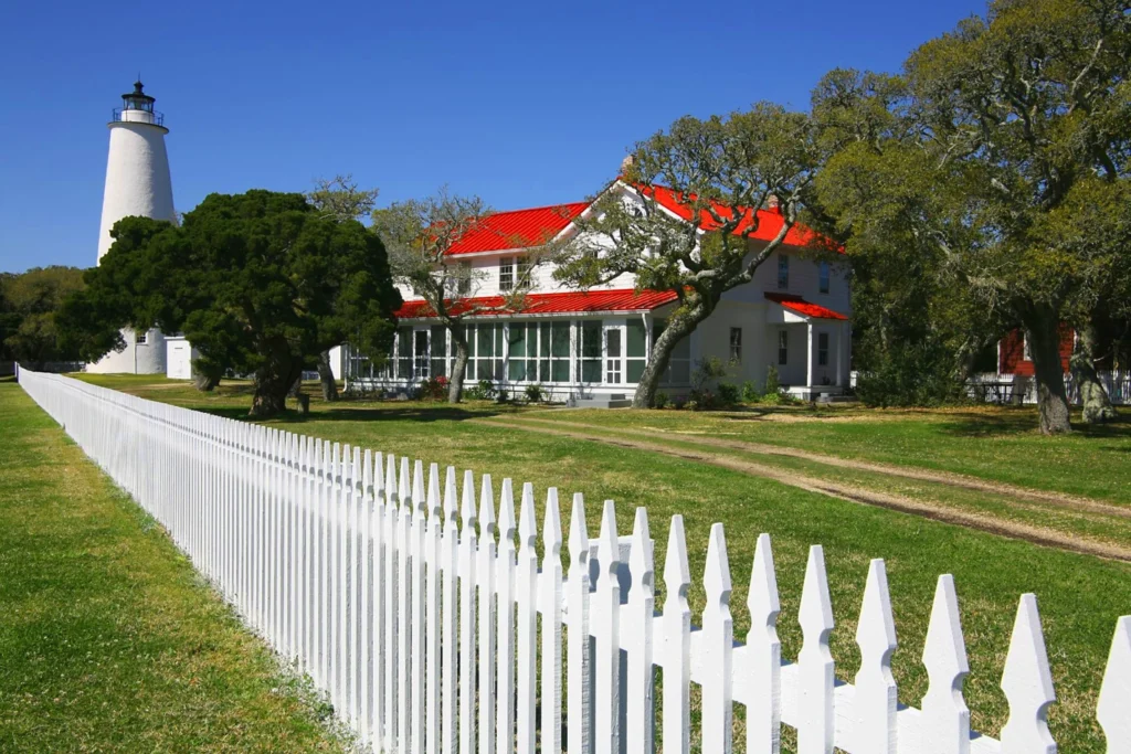 White Ocracoke Lighthouse standing beside a historic white two-story house with a bright red metal roof, framed by a white picket fence in the foreground, lush green grass, and mature trees under a clear blue sky