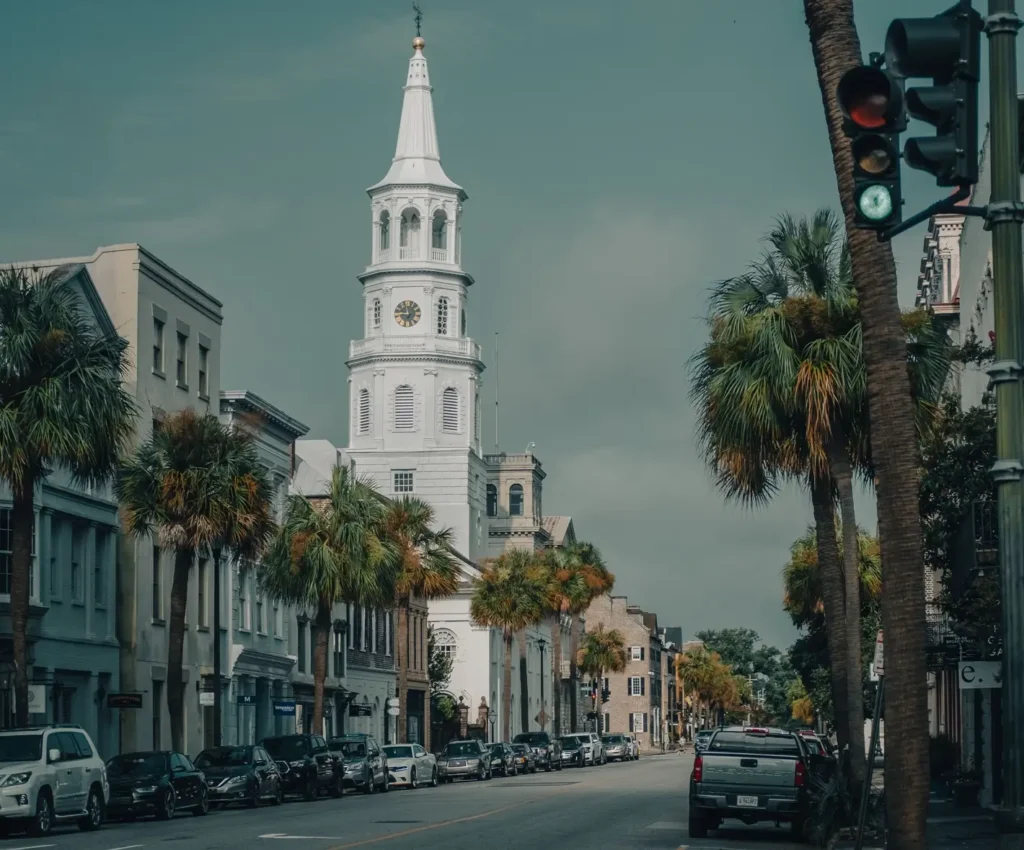 Street scene featuring the iconic white steeple of St. Philip's Church rising above palm trees and historic buildings in Charleston, with parked cars and traffic lights in the foreground.