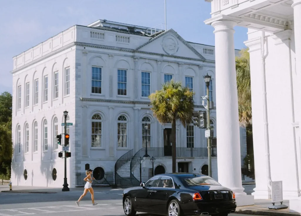 A woman jogging past the grand white classical-style building of the Charleston County Courthouse, with a black Rolls-Royce parked in front and large columns on the right.