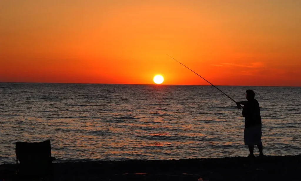 Silhouette of a person standing on a beach at sunset, fishing with a long rod over the ocean. The bright orange sun sits low on the horizon, casting a vibrant glow across the sky and water, with a beach chair visible on the sand in the foreground.