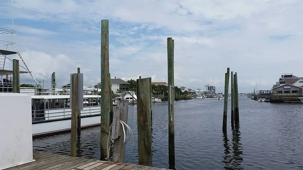View from a wooden dock looking out over a calm marina waterway lined with wooden pilings and ropes. Boats and waterfront buildings are visible in the background under a partly cloudy blue sky.