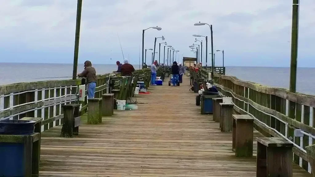 Long wooden fishing pier extending over the ocean, with several people fishing along the railings. The pier features lampposts, benches, and trash bins, stretching into the distance under an overcast sky.
