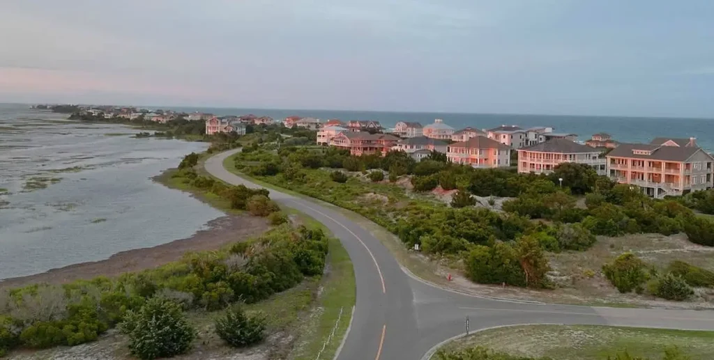 Aerial view of a coastal residential area at twilight, featuring large beach houses along a curving road that borders a marshy inlet on the left and the open ocean in the distance.
