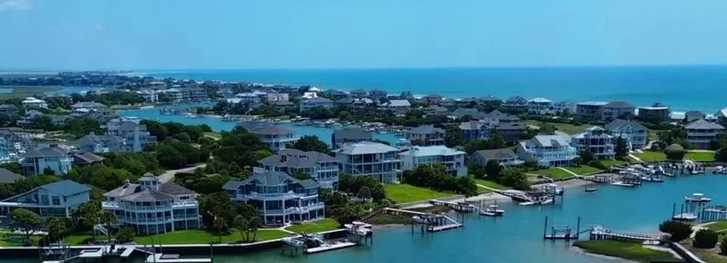 Panoramic aerial view of a waterfront residential community on a clear sunny day, showing numerous waterfront homes, private docks with boats, canals, and the blue ocean stretching to the horizon.