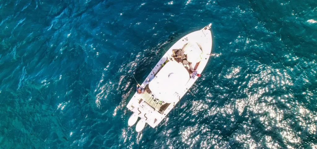 Aerial view of a white center console fishing boat with several people on board, surrounded by vibrant turquoise blue ocean water.