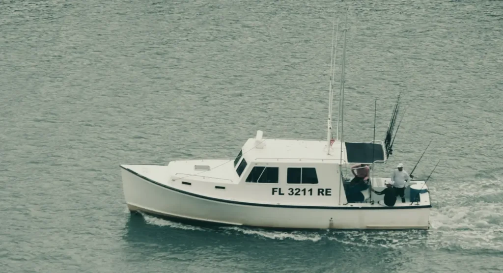 A white center-console fishing boat with registration FL 3211 RE cruising through greenish water. Two people are visible on deck, one in a white shirt and hat near the stern, with multiple fishing rods rigged up. The boat is moving and leaving a wake.