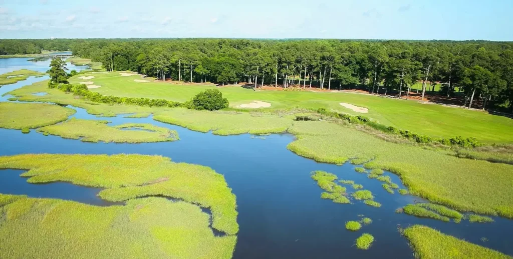 Aerial view of a stunning coastal golf course surrounded by expansive salt marshes, winding waterways, and dense pine forest under a bright blue sky.