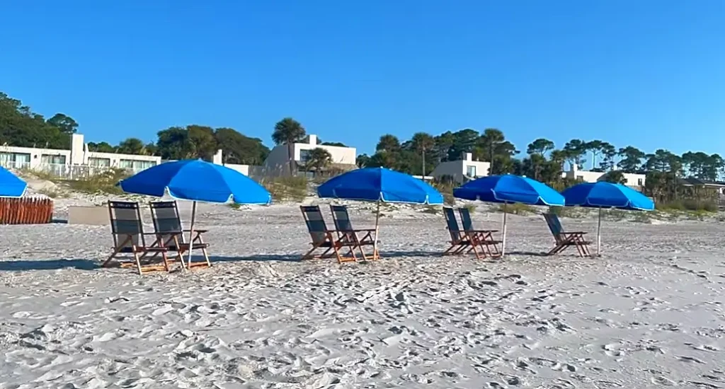Peaceful beach scene featuring rows of vibrant blue umbrellas shading wooden lounge chairs on soft white sand. Footprints are visible across the beach, with coastal homes and trees along the dunes under a bright, cloudless sky.
