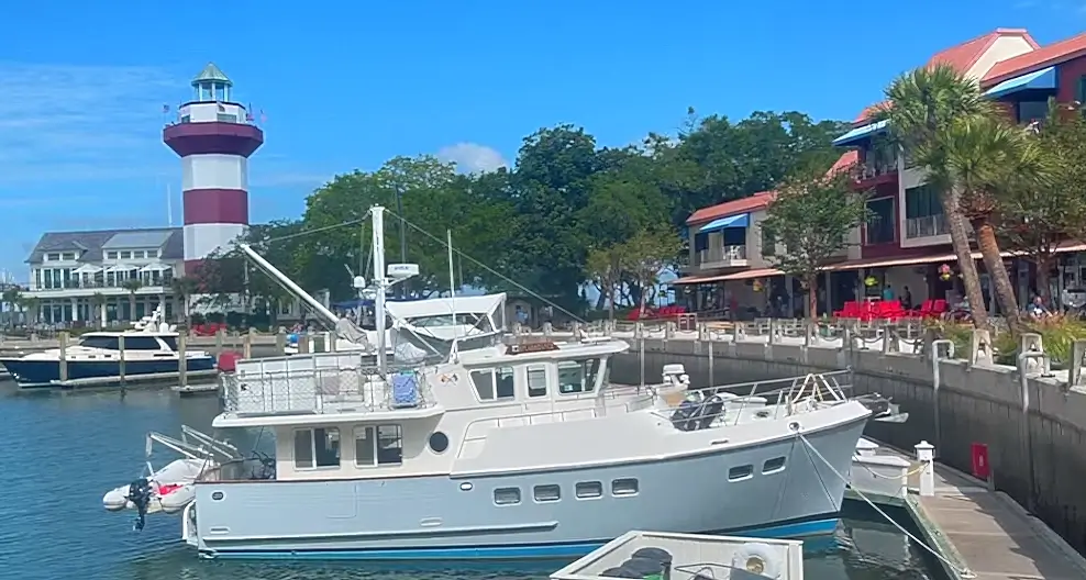A large white cabin cruiser with blue trim is docked in the foreground of a sunny coastal hilton head island marina. In the background stands a red and white striped lighthouse, surrounded by waterfront restaurants, palm trees, and other boats on a bright clear day.