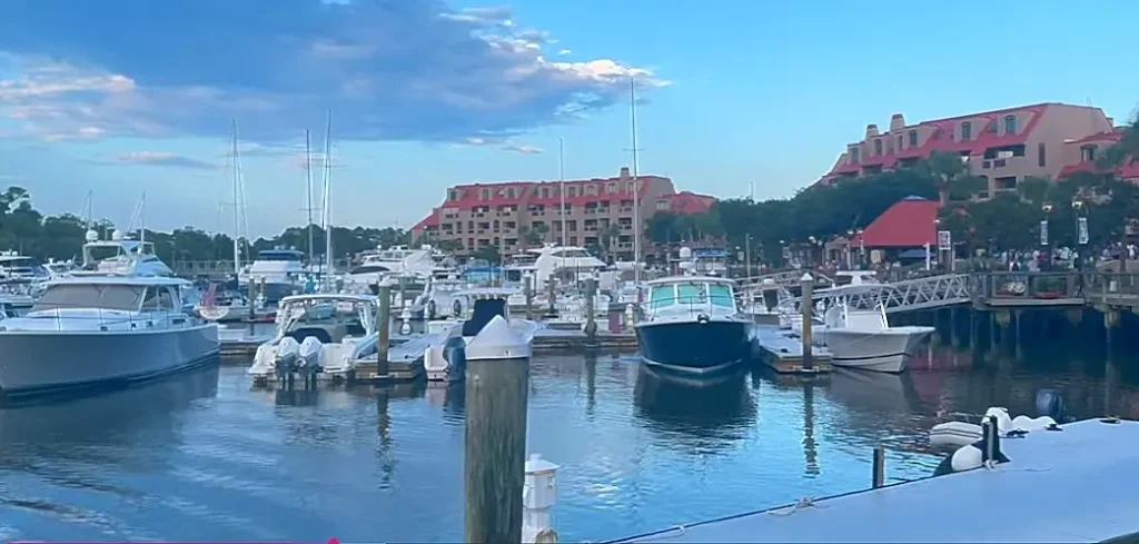 Wide view of a hilton head island harbor filled with motor yachts and sailboats moored at docks. A prominent beige building with red tile roofs overlooks the water, with people strolling on the boardwalk on a bright, sunny day.