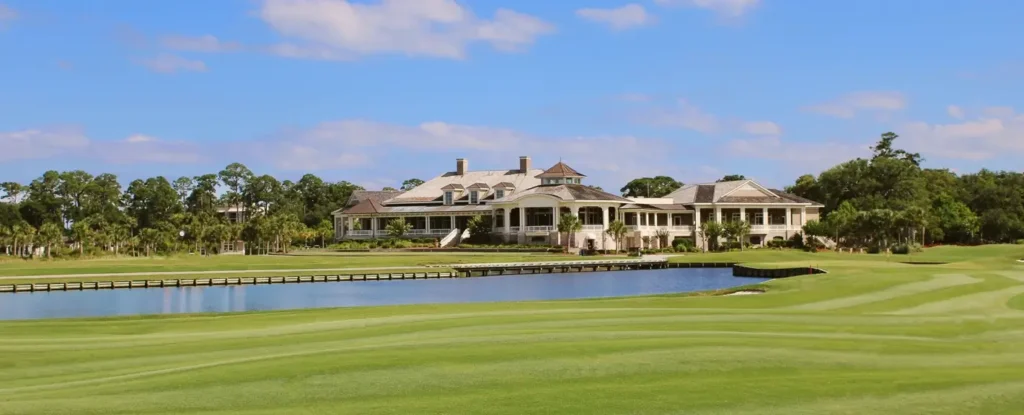 Beautiful waterfront golf course clubhouse with manicured fairways and a large pond in the foreground under a bright blue sky with scattered clouds.