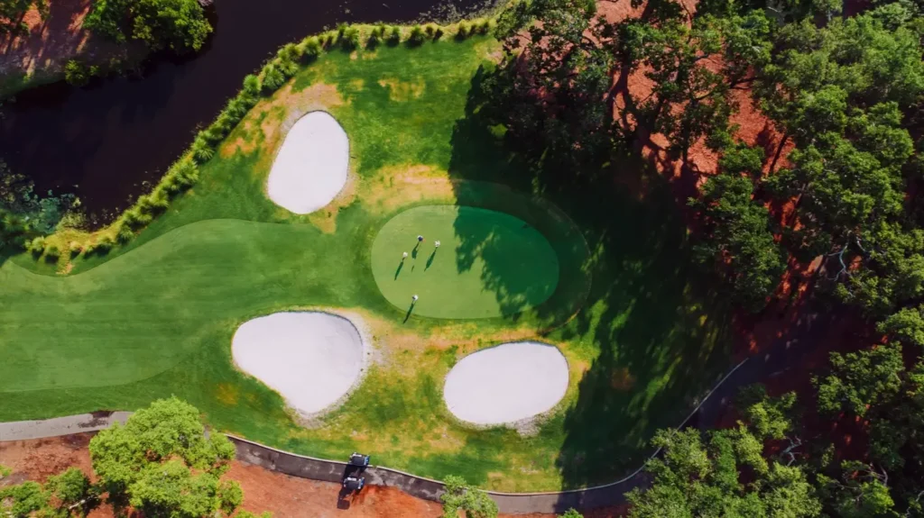 Aerial view of a golf green surrounded by three large sand bunkers, with several golfers putting. The green is bordered by water, trees, and a reddish dirt path with a golf cart below.