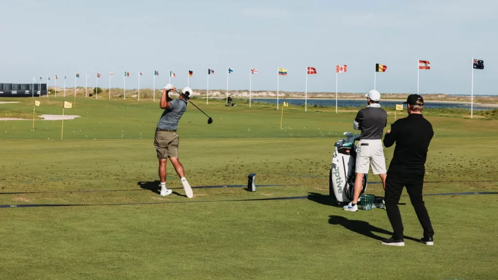 Golfer mid-swing on a driving range with international flags lining the range, overlooking the ocean and coastal dunes.