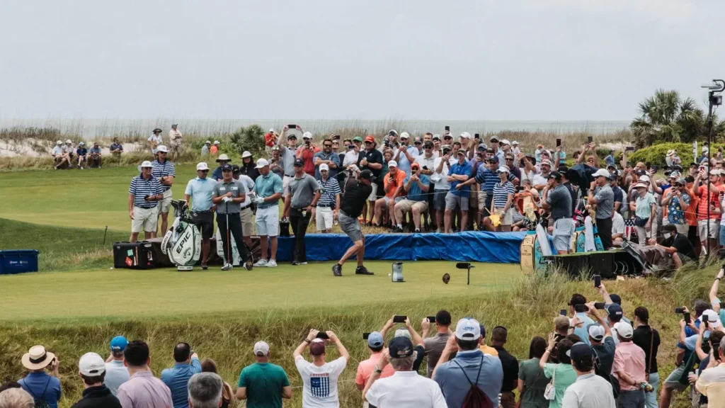 A professional golfer teeing off on a coastal course as a large gallery of spectators watches intently, with the ocean visible in the background during a PGA Tour event.
