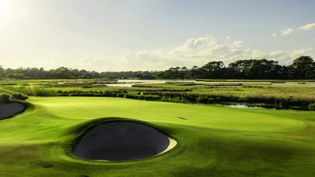Sunny wide view of a pristine golf green with a prominent sand bunker in the foreground. Marshland, water hazards, and distant trees frame the hole under a bright blue sky.