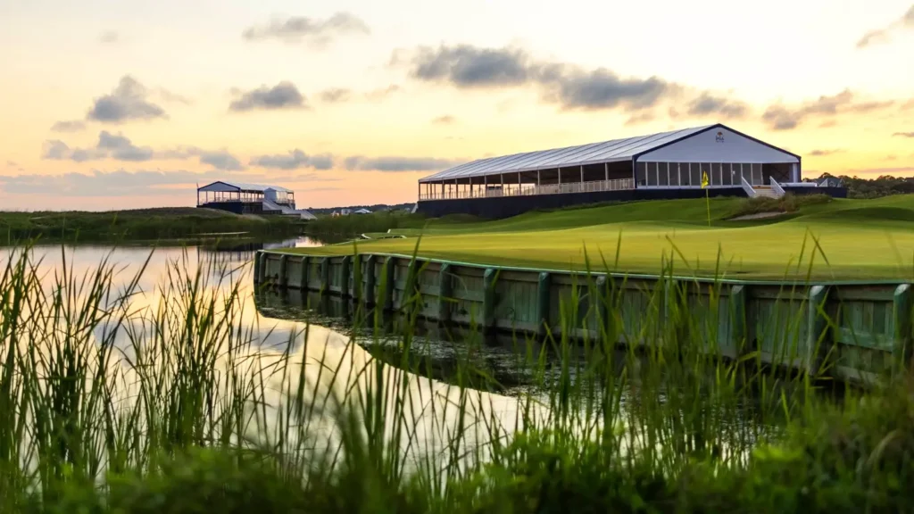 Golden sunset over a golf course with a large modern clubhouse and pavilion in the background. Reeds and calm water are in the foreground, with a green and flag visible on the right.
