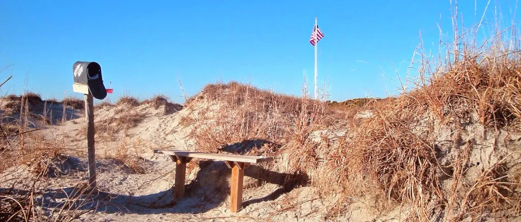 Black mailbox and simple wooden bench on a sandy path through dry dunes, with an American flag flying on a pole in the background under a clear blue sky.