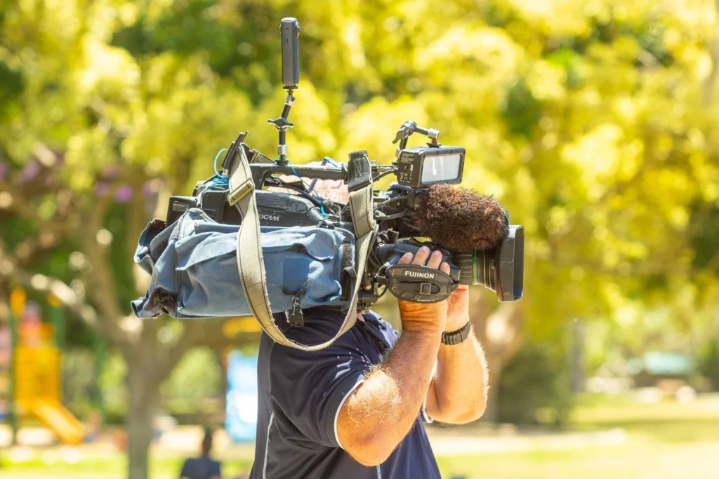 A cameraman from behind carrying a heavy professional shoulder-mounted video camera with a large furry windscreen microphone, filming outdoors in a sunny park with green trees in the background.