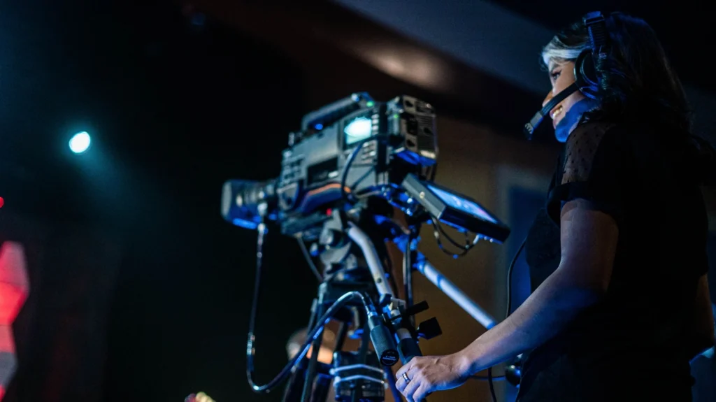 A woman with dark hair and white highlights wearing a professional headset with microphone, smiling while operating a large broadcast video camera on a tripod in a dark studio with dramatic blue lighting.