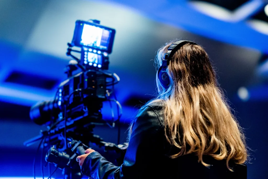View from behind of a woman with long blonde hair wearing blue headphones, operating a large professional video camera in a dimly lit blue studio environment.