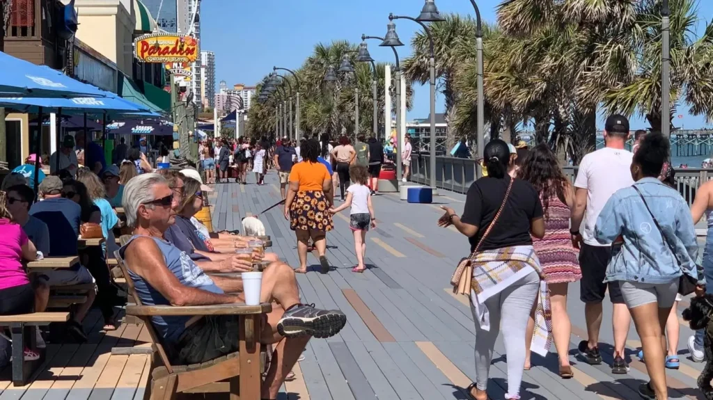 Crowds enjoying the Myrtle Beach Boardwalk with restaurants, oceanfront attractions, palm trees, and beach vacation atmosphere.