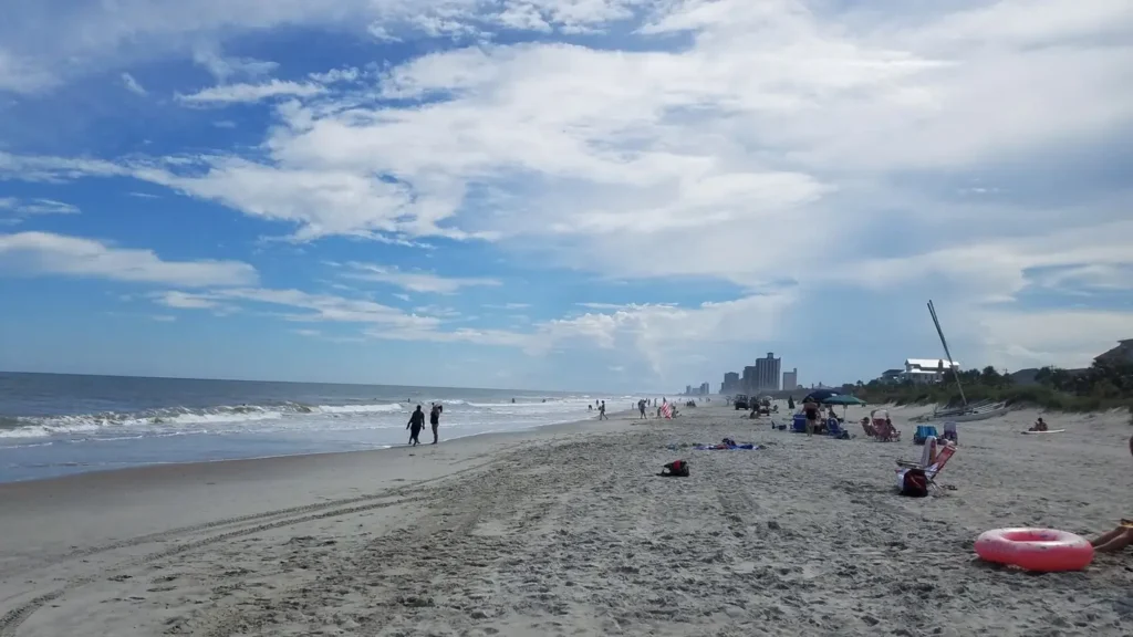 Wide shot of myrtle beach on a sunny day with scattered clouds in the blue sky. Gentle waves lap the shoreline on the left, where a couple is walking. Various beachgoers, umbrellas, and chairs are spread across the sand to the right, with high-rise coastal buildings visible in the distant background.