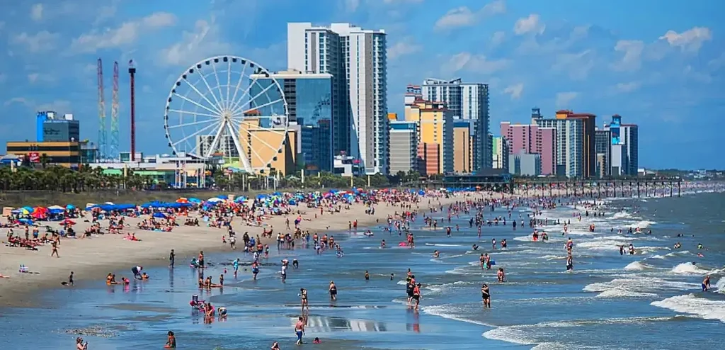 An expansive myrtle beach beachfront scene bustling with numerous people relaxing on the sand and wading in the shallow ocean surf under a partly cloudy sky. In the background, a coastal city skyline features a prominent white Ferris wheel alongside several high-rise hotels and resort buildings.