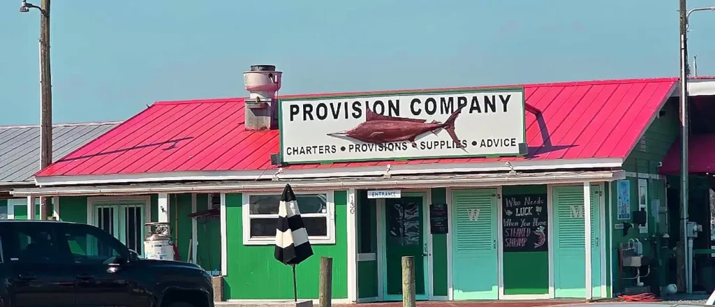 A sunny daytime view of the Provision Company storefront, a vibrant single-story building with bright green walls and a bright red metal roof. A large white sign on the roof features bold text reading 'PROVISION COMPANY' alongside a graphic of a marlin fish, with 'CHARTERS • PROVISIONS • SUPPLIES • ADVICE' below it. A black pickup truck is parked in the foreground, and various smaller signs adorn the entrance area.