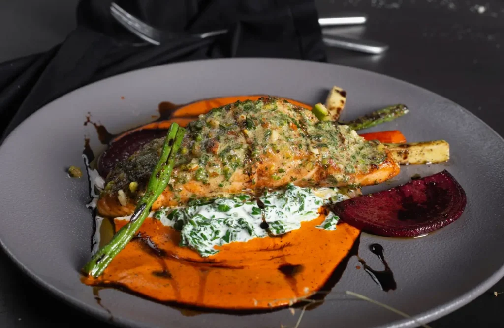A beautifully plated gourmet salmon fillet on a dark gray plate. The salmon is topped with a vibrant green herb crust and sits on a bright orange sweet potato purée. It's accompanied by grilled asparagus, roasted beets, and a dollop of creamy spinach sauce, all elegantly arranged with a dark balsamic glaze drizzle.