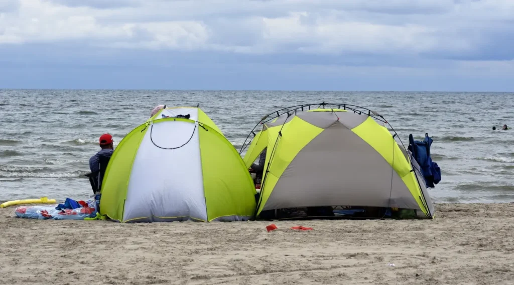 Two bright lime green camping tents set up on a sandy beach by the ocean under a cloudy sky. A person in a red cap sits in a chair beside the smaller tent, with beach gear scattered around and people swimming in the distance.