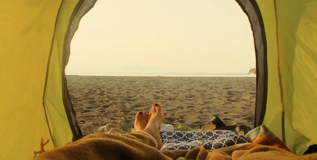 View from inside a yellow tent looking out onto a sandy beach and calm ocean. A person’s bare feet with black nail polish are relaxed in the foreground on a blanket, capturing a peaceful beach camping moment.