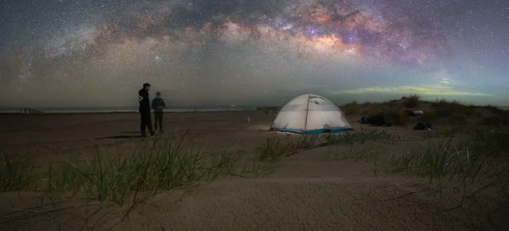 Two people standing on a sandy beach dune at night, gazing at a breathtaking starry sky filled with the colorful Milky Way. A illuminated white and blue camping tent is pitched nearby, with the ocean visible on the horizon.