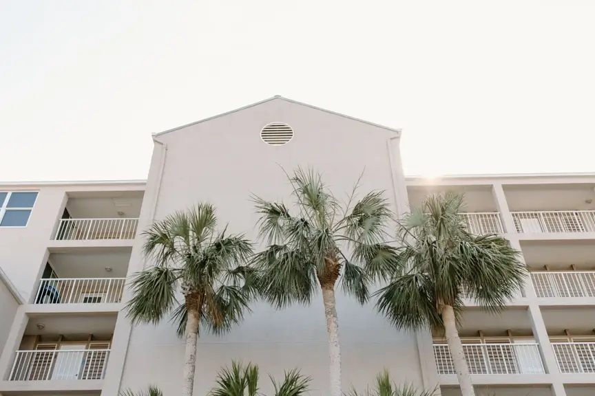 Low-angle view of a modern white multi-story vacation rental building with multiple balconies and white railings, framed by tall palm trees in the foreground against a bright, clear sky.
