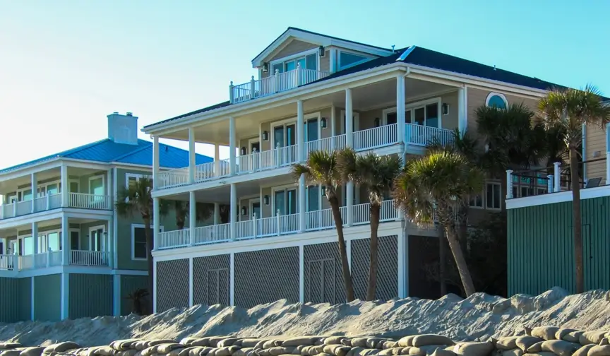 Large oceanfront vacation rental homes with wraparound porches and elevated foundations stand behind a sand dune and rock erosion barrier along a sunny Carolina beach, with palm trees swaying under a clear blue sky.
