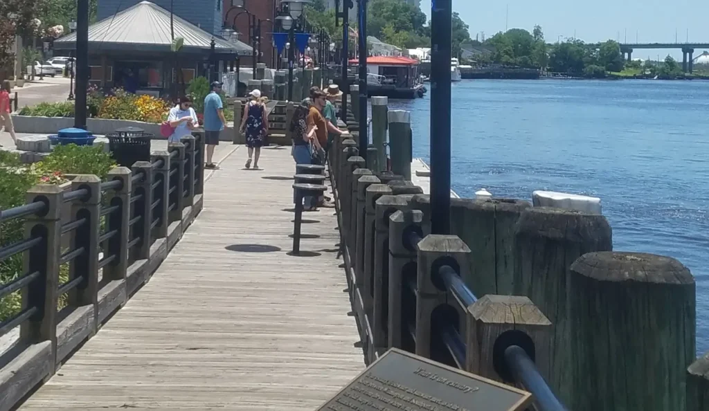 A sunny day on a wooden boardwalk along a waterfront. Several people are walking and standing along the pier, with shops, flower beds, and lampposts on the left and calm blue water on the right. Boats are visible in the distance, along with a large bridge on the horizon. A historical plaque is in the foreground.