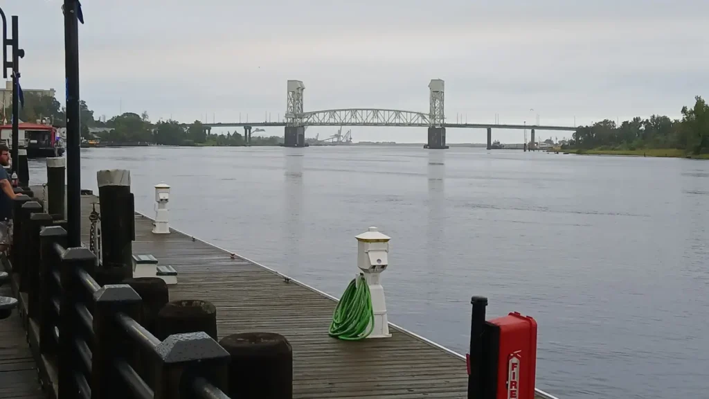A wooden pier overlooking a wide, calm river or bay under an overcast sky. A prominent large steel lift bridge spans the water in the distance. A green hose is coiled on a white post and a red fire equipment box are visible on the dock in the foreground.