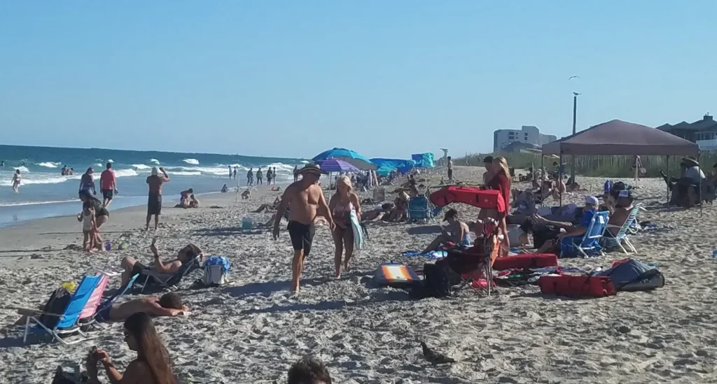 A lively, crowded beach on a bright sunny day. People are sunbathing, walking, and swimming in the ocean waves. The sand is filled with colorful beach chairs, umbrellas, and beachgoers. Buildings and dunes are visible in the background under a clear blue sky.