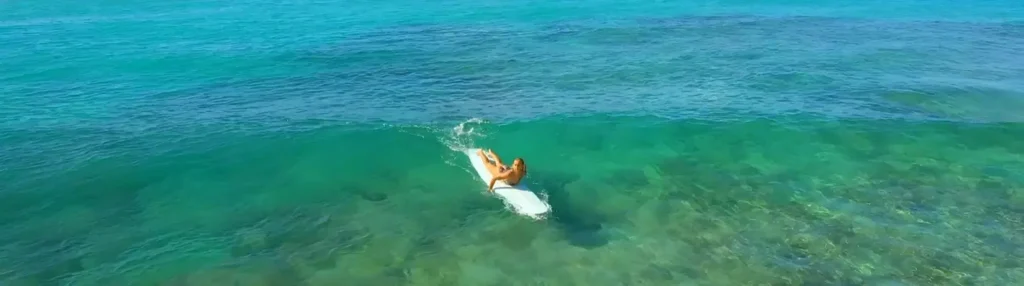 Aerial view of a person lying on a white surfboard in clear turquoise tropical water, riding a gentle wave over a visible seabed.