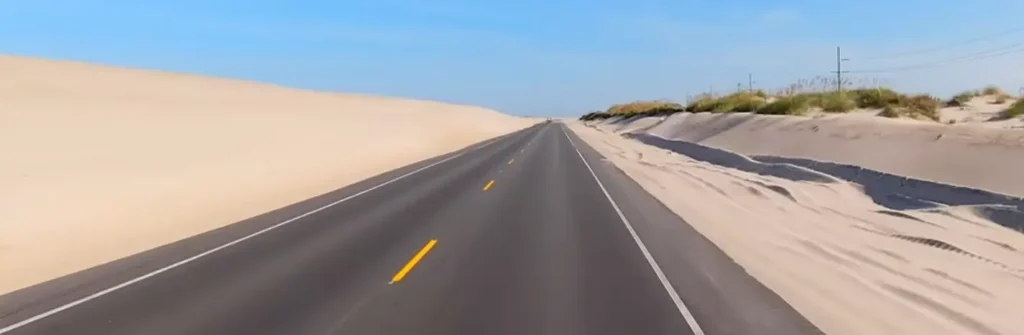 A dramatic road cutting directly between towering sand dunes on the left and lower vegetated dunes on the right, creating a corridor effect under clear skies.