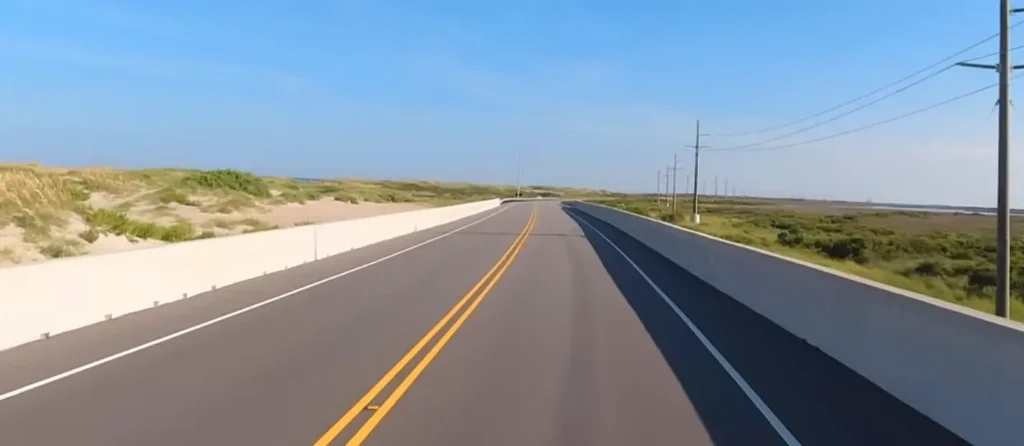 A straight two-lane road flanked by concrete barriers, with sandy dunes and coastal vegetation on both sides under a bright blue sky.