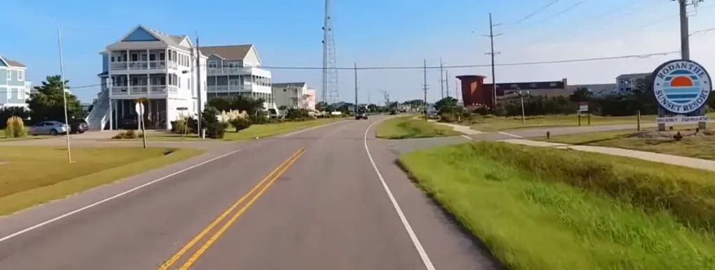 A coastal town street lined with large beach houses, utility poles, and a prominent “Rodanthe Sunset Resort” sign on the right under a clear blue sky.