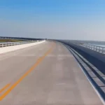 A long, elevated concrete bridge stretches straight ahead over calm water on a bright sunny day, with marshland visible on the left and open water on the right.