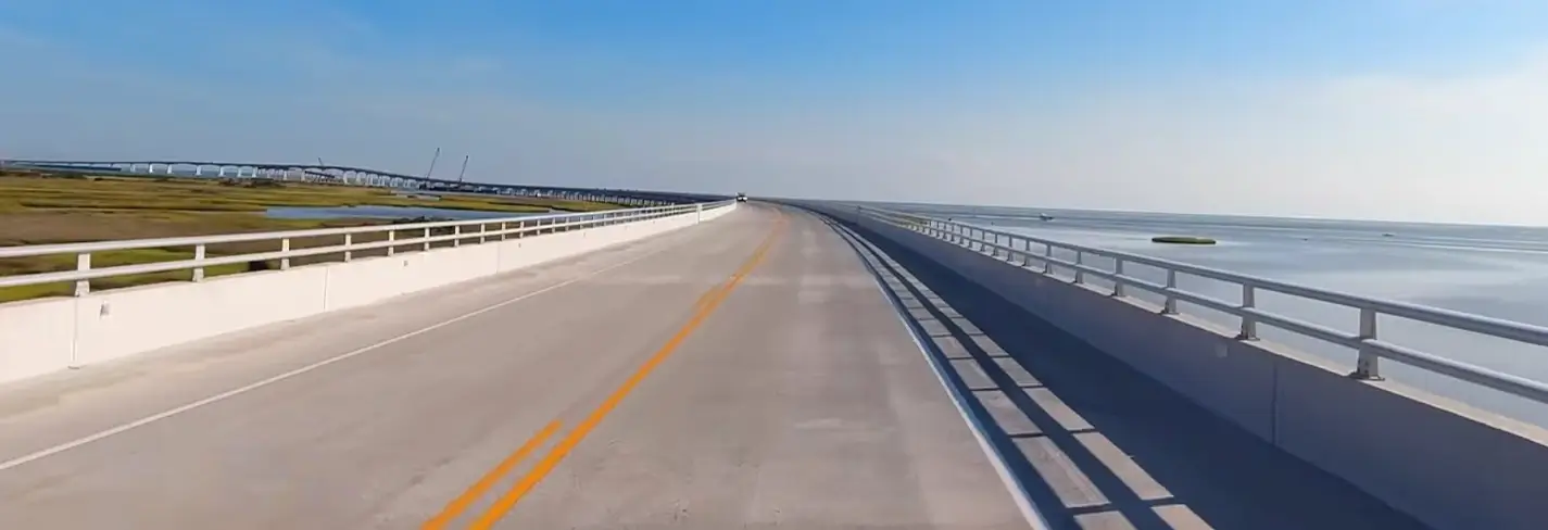 A long, elevated concrete bridge stretches straight ahead over calm water on a bright sunny day, with marshland visible on the left and open water on the right.
