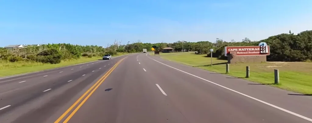 A paved two-lane road curves gently under a clear blue sky toward the entrance sign for Cape Hatteras National Seashore, with a silver car approaching and grassy coastal vegetation lining the sides.